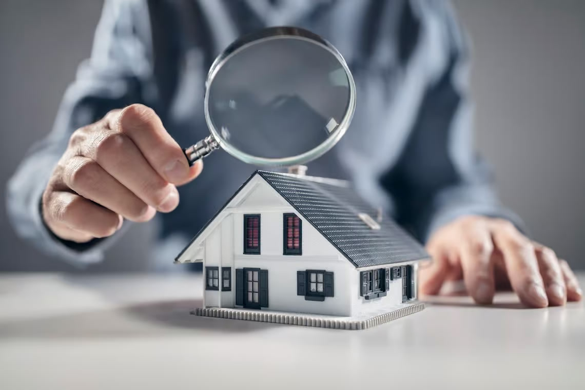 Hand holding a magnifying glass over a small white model house, representing a builder-realtor’s close look at a Kirkland home